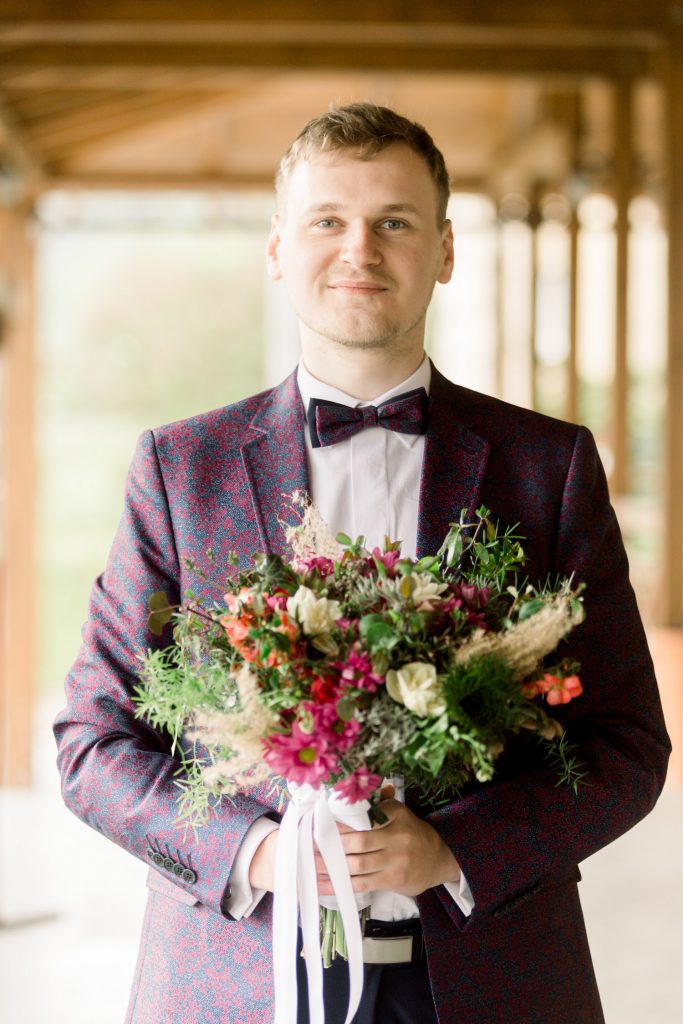 Ženích v bordovom obleku so svadobnou kyticou čaká na nevestu a ich first look